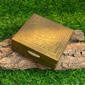 Pode incluir: Uma pequena caixa quadrada de latão com um design gravado intrincado de folhas e flores. A caixa tem uma tampa com dobradiças e uma trava de latão.