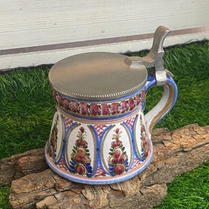 Peut inclure: Une chope à bière en céramique avec un couvercle métallique à charnière. La chope présente un motif floral coloré en rouge, bleu et or. La poignée est incurvée et le couvercle est fixé avec une charnière métallique. La chope repose sur un morceau de bois.