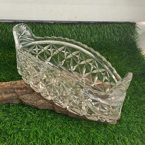May include: A clear, cut-glass serving dish with a decorative starburst pattern. The dish has a curved, boat-like shape and sits on a piece of wood. The edges of the dish are scalloped. The dish is displayed on a bed of green artificial grass.