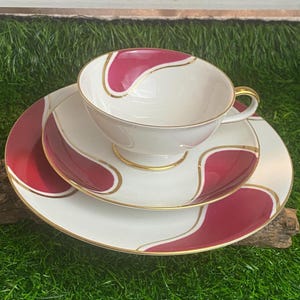 Könnte beinhalten: Ein weißes Tee-Set mit rotem und goldenem Wirbelmuster. Die Tasse hat einen Henkel und die Untertasse einen gewellten Rand.