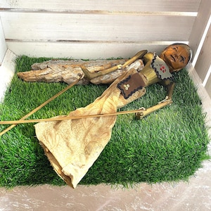 Könnte beinhalten: Eine Vintage-Holzpuppe mit bemaltem Gesicht und gegliederten Gliedmaßen, bekleidet mit einem goldfarbenen Stoffmuster. Die Puppe liegt auf einem Bett aus grünem Kunstrasen, mit einem Holzstück im Hintergrund. Die Arme der Puppe sind ausgestreckt und sie hat ein detailliertes Kostüm.