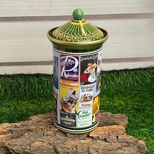May include: A cylindrical tin with a green lid and a green knob. The tin is decorated with vintage advertisements in various colors. The lid has a green and gold filigree design. The tin is sitting on a piece of wood.