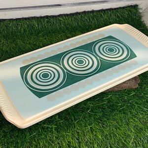Puede incluir: Bandeja rectangular para servir con borde color crema e interior azul claro. La bandeja presenta un diseño de círculos concéntricos verdes y blancos. La bandeja está colocada sobre un lecho de césped artificial verde.