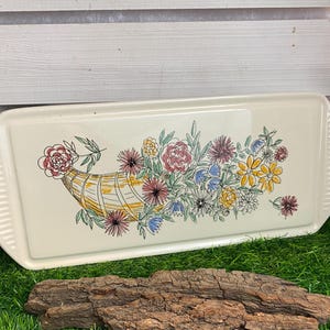 Könnte beinhalten: Ein weißes Keramik-Serviertablett mit einem Blumendesign, das eine Füllhorn zeigt, das mit bunten Blumen überquillt. Das Tablett hat einen erhöhten Rand und eine leicht strukturierte Oberfläche.