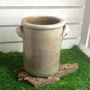 Könnte beinhalten: Ein zylindrischer Steinguttopf mit einer gedämpften beige-grauen Oberfläche und einem glatten, abgerundeten Rand. Das Gefäß verfügt über zwei kleine, geschwungene Seitengriffe am oberen Teil und weist eine verwitterte, erdige Textur auf.