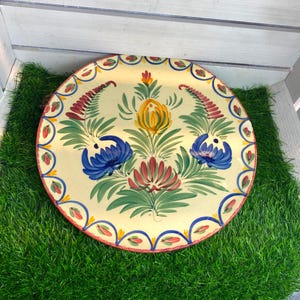 Könnte beinhalten: Ein runder Keramikteller mit cremefarbener Basis und handgemaltem Blumenmuster. In der Mitte befindet sich ein Arrangement aus gelben, blauen und roten Blüten mit grünen Blättern. Der Rand ist mit einem wiederkehrenden blau-roten Blütenblattmuster verziert.