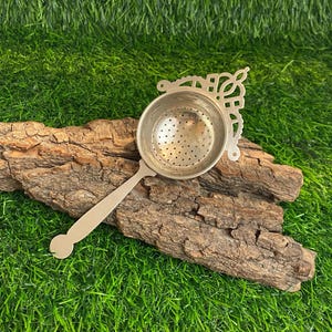 Op de afbeelding: Een zilveren thee-ei met een decoratief handvat en een geperforeerde kom. Het thee-ei ligt op een stuk hout tegen een achtergrond van groen gras. De handgreep heeft een afgerond uiteinde.