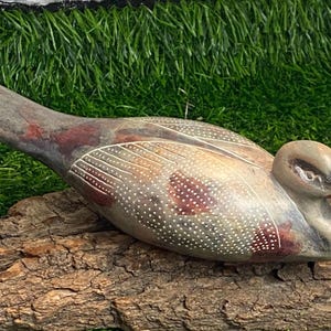 Könnte beinhalten: Eine handgeschnitzte Steinskulptur eines Enten mit einer gesprenkelten Oberfläche in Grau, Braun und Weiß. Die Ente liegt auf einem Stück verwittertem Holz.