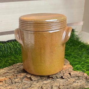 Könnte beinhalten: Ein brauner Keramiktopf mit Deckel und zwei seitlichen Griffen. Der Topf hat eine zylindrische Form und eine strukturierte Oberfläche. Der Deckel hat horizontale Rillen. Der Topf wird auf einem Holzstück ausgestellt.