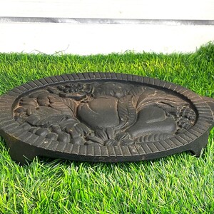 Puede incluir: Un salvamanteles ovalado de hierro fundido negro con un diseño en relieve de frutas variadas, incluyendo peras y uvas. La pieza presenta un borde texturizado, dos pequeñas asas laterales y patas cortas para elevarlo de la superficie.