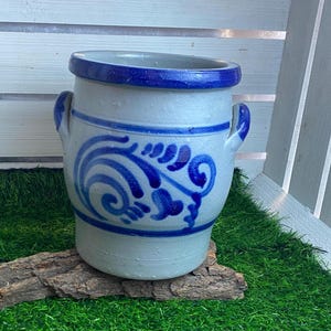 Op de afbeelding: Een witte keramische pot met een blauwe glazuur en een blauw bloemenontwerp. De pot heeft twee handvatten en een brede opening.