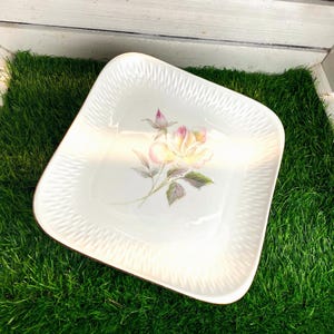 Puede incluir: Un plato cuadrado de cerámica blanca con una ilustración central de una rosa de color amarillo pálido y rosa con hojas verdes. El borde del plato tiene un patrón texturizado en relieve y un fino borde marrón que enmarca el diseño floral.