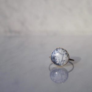 May include: A ring featuring a circular, white stone with black dendritic inclusions set in a polished metal band. The stone has a smooth, domed surface with a distinct pattern of dark, branching, fern-like markings across the white base.