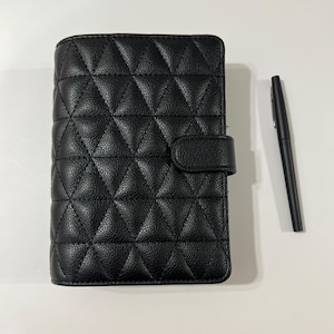 Könnte beinhalten: Schwarzes, gestepptes Lederjournal mit Klappenverschluss und schwarzem Stift. Das Journal hat ein Rautenmuster. Der Stift ist schwarz mit silberner Spitze. Das Journal ist ca. 15 cm hoch und 10 cm breit.