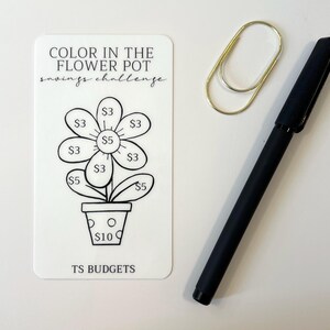 May include: A white savings challenge card with a flower pot design and the text "COLOR IN THE FLOWER POT savings challenge." A gold paperclip and a black pen are next to the card. The flower has amounts from $3 to $10.