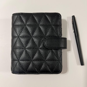 May include: Black quilted leather planner with a snap closure. The planner has a diamond pattern and a matching black pen is next to it. The planner is a compact size, perfect for carrying in a bag or purse.