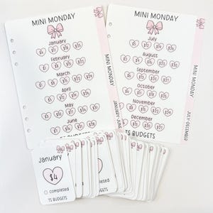 May include: Two white planner inserts and a stack of small cards featuring the text MINI MONDAY and TS BUDGETS. Each page lists months from January to December with heart-shaped icons containing dollar amounts and a pink bow illustration at the top.