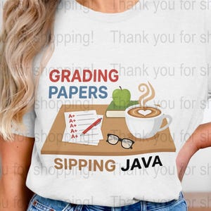 Può includere: T-shirt bianca con grafica di una scrivania con compiti in classe, una tazza di caffè con un cuore, una mela e occhiali. Il testo recita "GRADING PAPERS SIPPING JAVA."