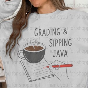 Peut inclure: Sweat-shirt blanc avec l'inscription "GRADING & SIPPING JAVA" en noir. Le motif comprend une tasse de café fumante, un bloc-notes et une main tenant un stylo rouge. Le sweat-shirt est de style décontracté.