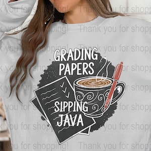 Peut inclure: Sweat-shirt blanc avec un motif graphique comportant les mots "GRADING PAPERS" et "SIPPING JAVA". Le motif comprend une tasse de café, un stylo et des papiers avec des coches. Un haut décontracté et confortable.