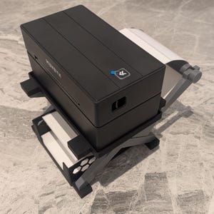 May include: A black rectangular thermal label printer with a white paper roll attached to the back. The device features a top-mounted power button with a blue indicator light and the brand name MUNBYN printed in white on the side of the housing.