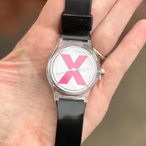May include: A clear plastic wristwatch with a black band. The watch face is white with a large pink "X" in the center. The watch has silver-colored hands and a silver-colored crown.