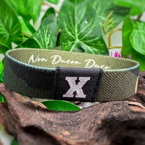 May include: A green and black camouflage wristband with the text "Non Ducon Duco" and a white letter "X" on a black patch.