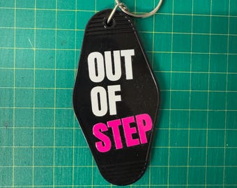 Può includere: Portachiavi nero in stile motel con le parole "OUT OF STEP" in lettere bianche e rosa. Il portachiavi ha un anello di metallo argentato attaccato in alto. Lo sfondo è una griglia verde.