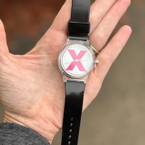 May include: A clear wristwatch with a black band. The watch face features a white background with a large pink "X" design. The watch is held in a hand, showcasing the watch's design and band.