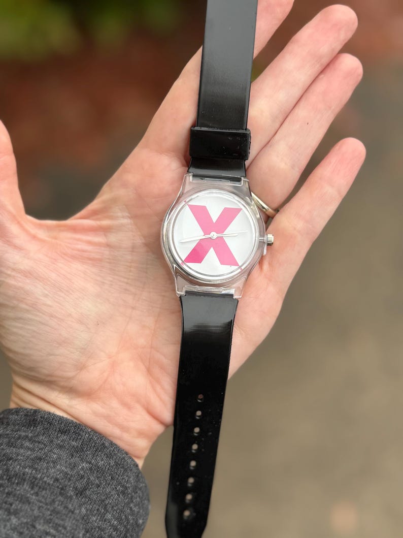 May include: A clear wristwatch with a black band. The watch face features a white background with a large pink "X" design. The watch is held in a hand, showcasing the watch's design and band.