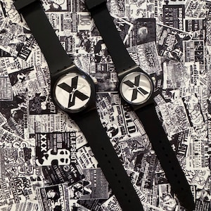 Può includere: Due orologi da polso neri con cinturini in silicone opaco e casse rotonde. I quadranti bianchi presentano un grande design a forma di X nera al centro, con lancette nere per ore, minuti e secondi. Gli orologi hanno fibbie in metallo argentato.