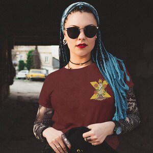 Könnte beinhalten: Eine Frau trägt ein weinrotes T-Shirt mit einem gelben, roten und grünen Grafikdesign. Die Grafik zeigt eine stilisierte Figur mit einer Krone und Blumen. Die Frau hat blaue Zöpfe und trägt eine Sonnenbrille.