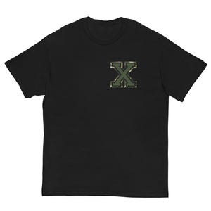 Op de afbeelding: Zwart T-shirt met een camouflage-patroon letter "X" patch op de linkerborst. De patch heeft tinten groen, bruin en bruin. Het shirt heeft korte mouwen en een ronde hals.