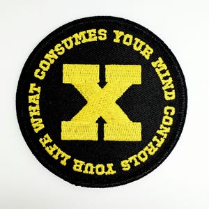 May include: A black and yellow embroidered patch with the text "What consumes your mind controls your life" and a large yellow "X" in the center.
