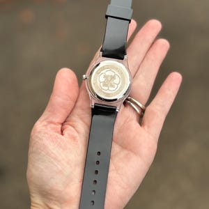 May include: A black wristwatch with a clear case and black band is held in a hand. The watch face is visible, featuring a gold-colored design. The watch band is made of a flexible material and has several holes for adjustment.