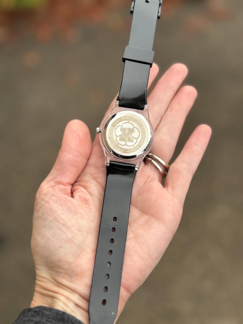 May include: A black wristwatch with a clear case and black band is held in a hand. The watch face is visible, featuring a gold-colored design. The watch band is made of a flexible material and has several holes for adjustment.