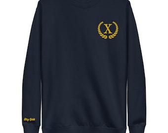May include: Navy blue sweatshirt with a gold embroidered "X" inside a laurel wreath on the chest. The left sleeve cuff has the text "Stay Gold" in gold. A casual, crew neck style.