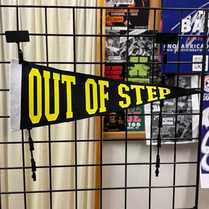 Peut inclure: Une bannière noire avec les mots "OUT OF STEP" en lettres jaunes. La bannière est affichée sur une grille métallique noire, avec des affiches de concert en arrière-plan. La bannière a un bord supérieur argenté et des glands noirs.