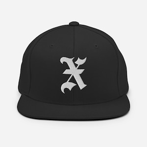 May include: Black baseball cap featuring a white embroidered gothic letter "K". The cap has a flat peak and a structured crown. A fashionable accessory for everyday wear.