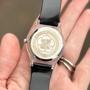 May include: A clear watch with a black band. The watch face features a silver emblem with the letter "X" at the center, surrounded by text. The watch is held by a person.