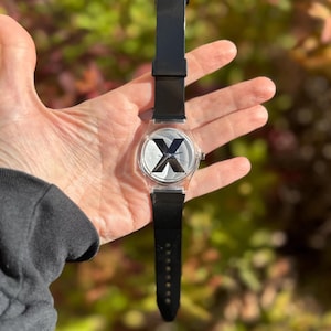 May include: A clear wristwatch with a black strap. The watch face features a silver background with a large black "X" design. The watch is held in a hand against a blurred background of green and brown.