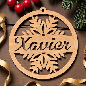 May include: A circular wooden ornament featuring a laser-cut snowflake design with the name Xavier engraved in a cursive script across the center. The ornament includes a small hole at the top for hanging and has a natural light brown wood finish.