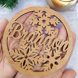 May include: A wooden Christmas ornament with a snowflake design and the name "Brandon" cut out. The ornament is light brown and has a rustic look.