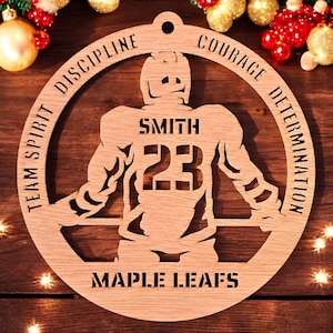 May include: A circular light brown wooden ornament featuring a laser-cut hockey player silhouette. The text reads SMITH 23, MAPLE LEAFS, TEAM SPIRIT, DISCIPLINE, COURAGE, and DETERMINATION. The design includes a small hole at the top for hanging.
