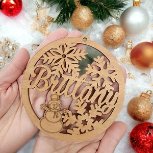 May include: A wooden Christmas ornament with a snowflake design and the name "Brittany" cut out. A snowman is also visible in the center of the ornament.