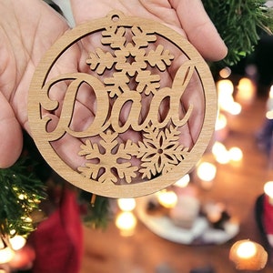 May include: A wooden Christmas ornament with a snowflake design and the word "Dad" in a cursive font.