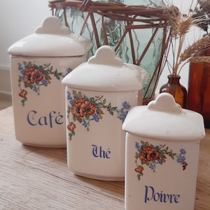 Puede incluir: Tres botes de cerámica blanca con diseños florales y letras azules. Los botes están etiquetados como "Cafe", "The" y "Poivre".