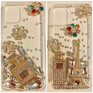 May include: Clear phone cases embellished with sparkling rhinestones and gold-toned accents. One case features a perfume bottle design with "PARFUM N°5" text, the other has a perfume bottle and Eiffel Tower design. Both cases have scattered rhinestones.