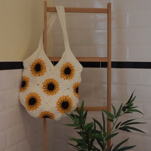 Puede incluir: Una bolsa de mano de crochet blanca con un patr&oacute;n de girasol. La bolsa est&aacute; colgada en un estante de madera contra una pared de azulejos blancos.