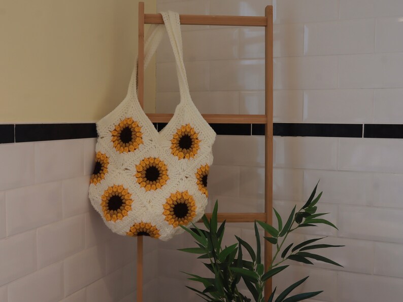 Puede incluir: Una bolsa de mano de crochet blanca con un patr&oacute;n de girasol. La bolsa est&aacute; colgada en un estante de madera contra una pared de azulejos blancos.